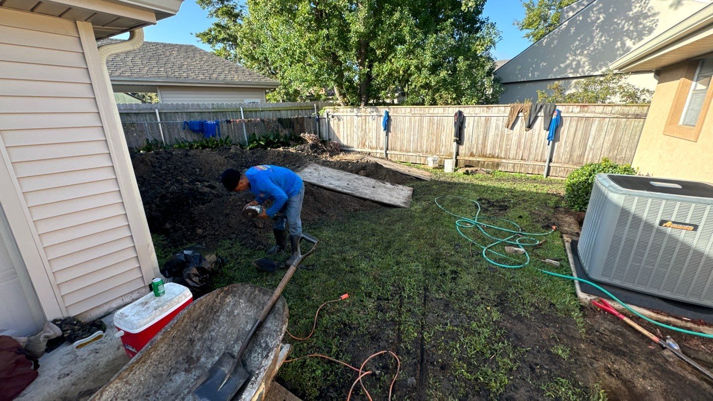 In New Orleans, a person is busy in their backyard, tackling repairs for a sinking foundation with a shovel. Amidst the wooden planks, garden hose, cooler, and air conditioning unit, freshly washed laundry sways gently on the fence in the vibrant Louisiana breeze.