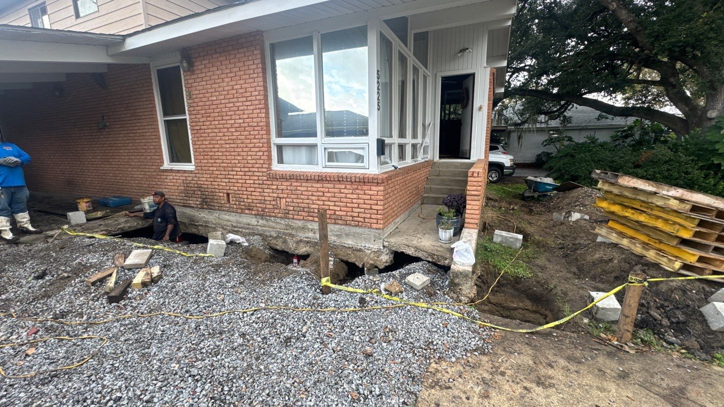 A house in St. Tammany Parish shows signs of foundation issues, with exposed earth, construction materials, and caution tape encircling the area as workers address foundation problems.