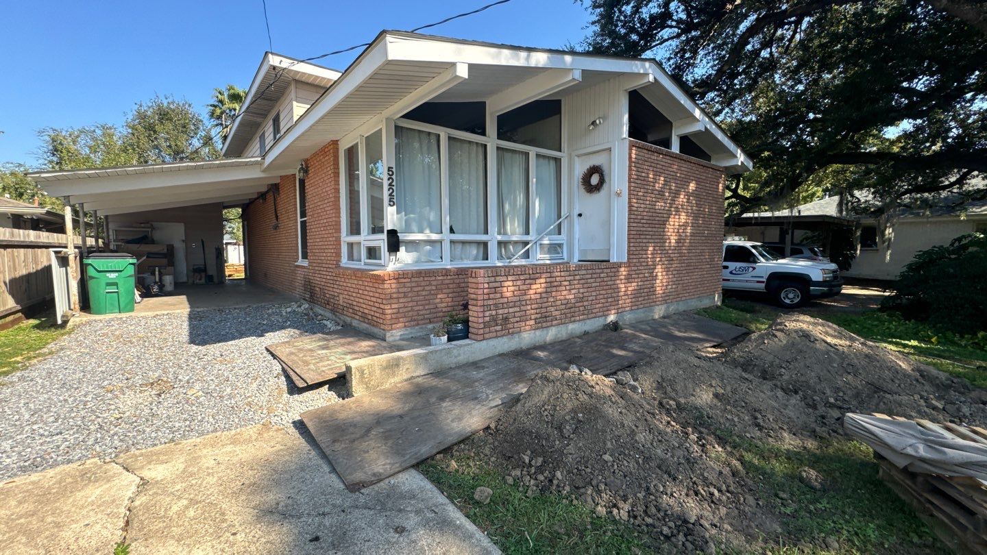 A brick house in St. Tammany Parish features a carport and a partially dug-up front yard, hinting at recent efforts to address foundation issues. A white truck is parked in the driveway, accompanied by a green trash bin near the sidewalk.