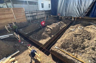 Two construction workers are diligently digging trenches for foundations at a Louisiana residential site. The area is marked with red string lines and surrounded by piles of dirt, while nearby, a building is covered with tarps to prevent any weather-related fail.