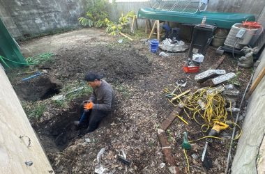 A man is diligently working in a Metairie, LA backyard, surrounded by tools, wires, and debris as he addresses foundation issues next to a large green tank.